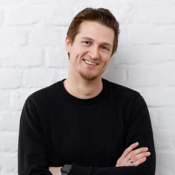 Junger Mann mit kurzen braunen Haaren lächelt in die Kamera, trägt schwarzen Pullover und Smartwatch, steht mit verschränkten Armen vor weißer Backsteinwand.